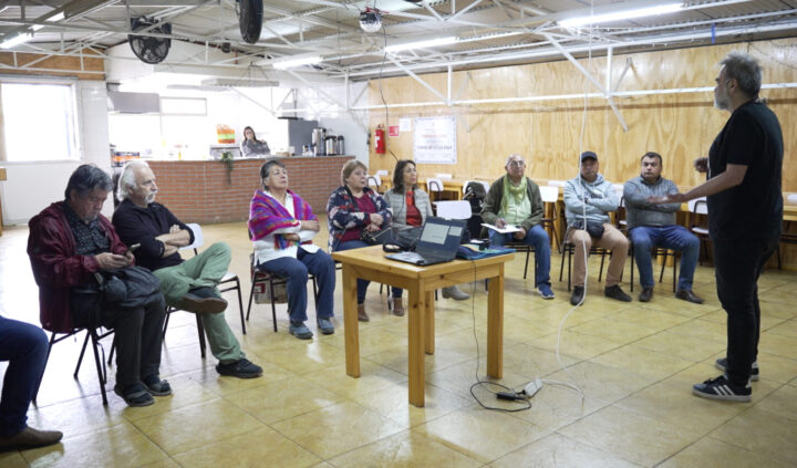 Un grupo de personas mayores participando en una sesión de capacitación cultural. Hay una presentación en una mesa al frente y un facilitador hablando. El ambiente es acogedor y la sala está decorada de manera sencilla.