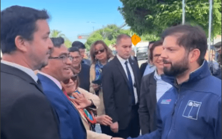 Presidente Gabriel Boric con chaqueta azul saluda a un hombre de traje. Multitud de personas observan en evento público al aire libre.