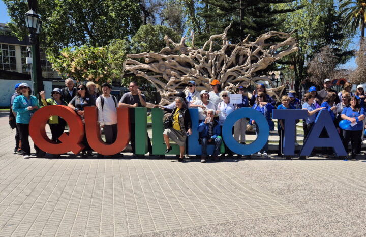 Grupo de personas sonrientes posando frente a las coloridas letras gigantes de Quillota. Fondo con árboles y escultura de raíces.