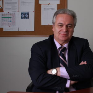 El autor de la columna, Juan Francisco Ortún, vestido con un traje oscuro y una camisa clara, sentado en un escritorio con las manos cruzadas y una expresión seria, frente a un fondo de documentos en un tablero de corcho.