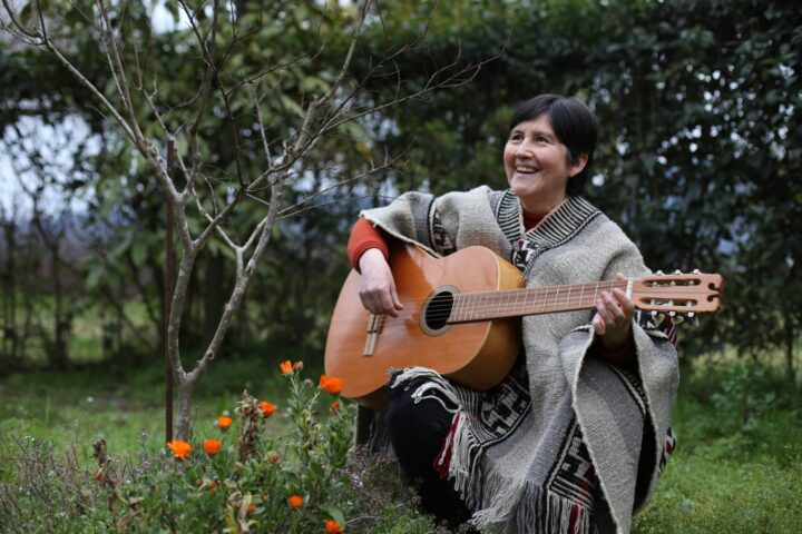 Mauricia Saavedra tocando una guitarra en un entorno rural, rodeada de flores y plantas.