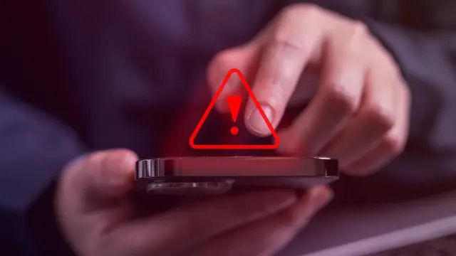 Una persona interactuando con un teléfono móvil que muestra un símbolo de advertencia en rojo, indicando peligro o alerta.